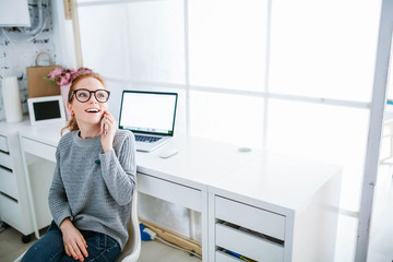 Wall Mural - Young beautiful woman with red hair, wearing glasses, working in the office, uses a laptop and mobile phone