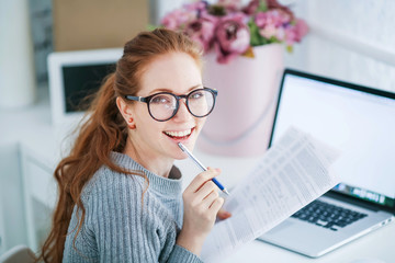 Wall Mural - Young beautiful woman with red hair, wearing glasses, working in the office, uses a laptop and mobile phone