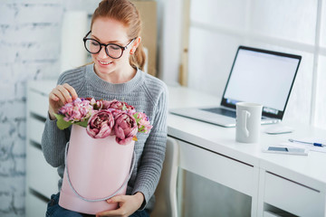 Wall Mural - Young beautiful woman with red hair, wearing glasses, working in the office, uses a laptop and mobile phone