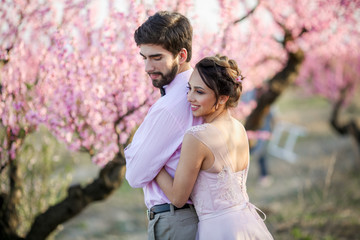 Newlyweds in love stand on the nature, against the background of wooden stakes, in sunny weather. Stylish groom embraces a beautiful bride in a lace dress in a green garden.