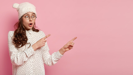 Wall Mural - Astonished dark haired woman with curly hair points aside, has shocked expression, wears hat with ears and casual shirt, shows copy space for your promotional content or advertisement. Look there