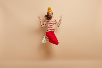 Isolated shot of happy overjoyed young woman jumps high, wears warm winter hat, jumper and red trousers, expresses happiness, isolated over beige background, celebrates victory. Emotions concept