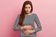 © wayhome.studio  - Indoor shot of displeased Caucasian woman suffers from period cramps, holds hands on belly, has pain in stomach, looks with upset expression, wears striped clothes, isolated over pink background