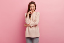 Photo Of Positive Pleasant Looking European Woman Keeps Hands Partly Crossed, Smiles Gently, Looks Directly At Camera, Wears Oversized Sweater, Poses Over Pink Background, Talks With Friend.