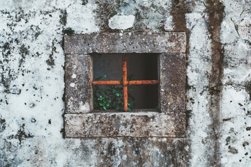 The fragment of an old grungy antique wall in Sintra, Portugal with a square stone window hole in the center with two rusty metal bars inside and a green plant, the facade of an abandoned house