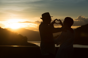 Silhouette of image of happy romantic senior couple making heart shape with hands outdoor at sunset  background