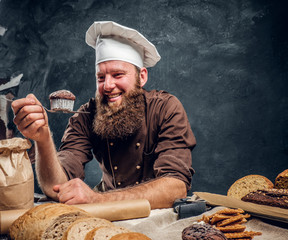 A bearded baker happy with his work, looking at his freshly made muffin while leaning on a table in a dark studio