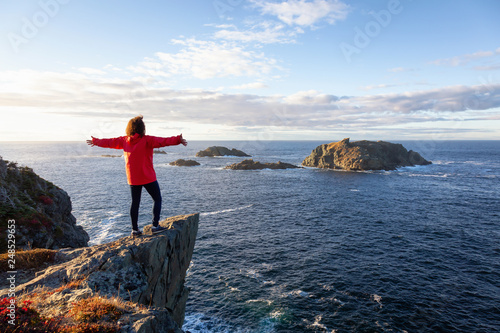 Fotomural Woman in red jacket is standing at the edge of a cliff with open arms and enjoying the beautiful ocean scenery