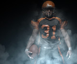 Fototapeta Sport - American football player on a dark background in smoke in black and orange equipment.