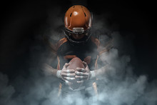 American Football Player On A Dark Background In Smoke In Black And Orange Equipment.