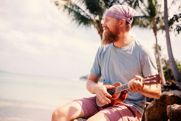 Young handsome redhead man with a beard playing a ukulele on a tropic beach, music, art, travel and vacations concept