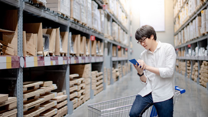 Wall Mural - Asian man shopper standing with trolley cart in warehouse inventory aisle checking his shopping list. Buying or purchasing factory goods. Shopaholic concept