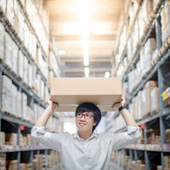 Wall Mural - Asian delivery man carrying cardboard box overhead in product shelves aisle in warehouse. Inventory picking and packing. Shopping lifestyle in department store. Buying or purchasing factory goods