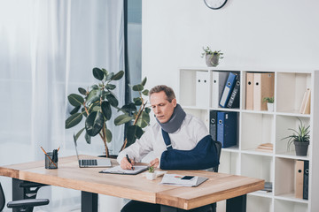 worker in neck brace and arm bandage sitting at table and carefully writing in office, compensation concept