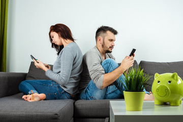 A man and a young woman, sitting on the couch and holding smartphones in their hands, turned away from each other. The concept of apathy with relationships, boredom, personal space, copy space.