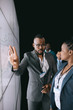 © JonoErasmus - Black african businessman looking at female colleague pointing to large video screen for a presentation during a conference.