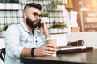 © foxyburrow - Young happy bearded man having mobile phone conversation with business partner, while sitting with laptop in cafe.
