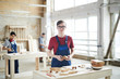 Content young carpenter wearing glasses and blue uniform standing at table and holding wooden piece while working with wood at modern factory