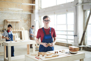 Wall Mural - Content young carpenter wearing glasses and blue uniform standing at table and holding wooden piece while working with wood at modern factory