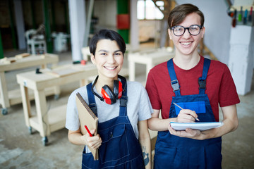 Cheerful satisfied young students in workwear standing in modern carpentry classroom and laughing while looking at camera and writing down information in sketchpad