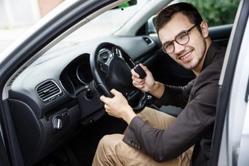 Wall Mural - Satisfied young driver is sitting at his car while looking at the camera. He is holding the keys at his right hand. His left hand is on the steering wheel.