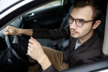 Wall Mural - Pensive young guy is looking through the window. He is sitting at his car. His right hand is on the steering wheel.