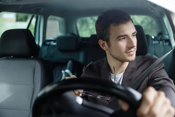 Wall Mural - Delighted young man is looking trough the window while driving his car. He is holding his glasses at his right hand. His left hand is on the steering wheel.