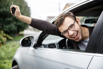 Wall Mural - Very happy young man is peeking from the car window while looking at the camera. He is holding the keys at his right hand. Lottery winner concept.