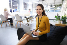 Positive Beautiful Young Woman With Badge Sitting In Comfortable Bean Bag And Smiling At Camera, While Typing On Laptop, She Working In Cozy Office