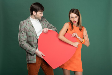 man looking at dissatisfied woman holding heart shaped paper card isolated on green