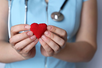 Health care, cardiology, woman doctor with stethoscope holding red knitted heart in hands. Concept of cardiologist, blood donation, charity, treatment of heart disease in the clinic