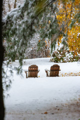 Two empty adirondack chairs on an early snowfall in the autumn.