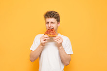 Handsome Young Man Bites A Piece Of Appetizing Pizza And Looks At The Camera On A Yellow Background. Boy In A White T-shirt Eats Fast Food, Pizza, Isolated On A Yellow Background. Copyspace