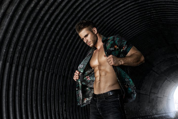 Muscular young man with beard on dark tunnel urban background. Fashion portrait of brutal strong muscle guy with modern trendy hairstyle. Model, fashion concept. Sexy naked torso, six pack abs.