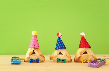 Purim celebration concept (jewish carnival holiday). Traditional hamantaschen cookies with cute clown hats and noisemaker over wooden table.