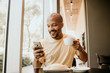 © KleverLevel - Happy cheerful African hipster holding mug, drinking fresh cappuccino, browsing internet and checking newsfeed on social media.Man using cell phone during coffee break at modern cafe