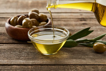 Pouring Oil From Bottle Into Glass Bowl, Bowl Of Olives, Olive Tree Branch And Olive On Wooden Surface