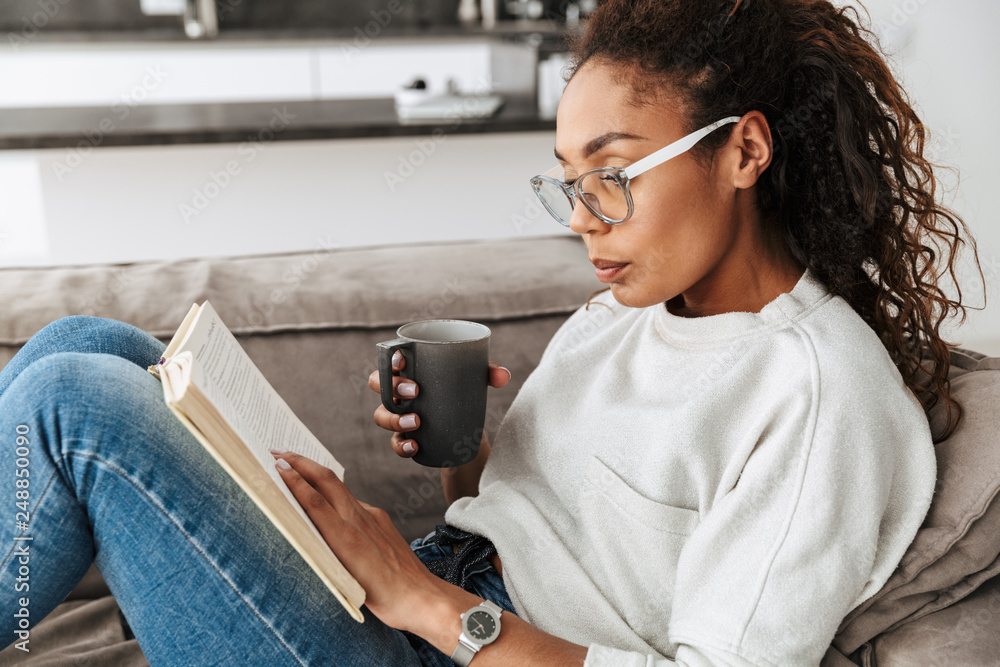 Image of smart african american girl reading book and drinking tea, while sitting on sofa in bright flat - obrazy, fototapety, plakaty 