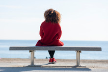 Rear View Of A Young Curly Woman Wearing Red Denim Jacket Sitting On A Bench While Looking Away To Horizon Over Sea