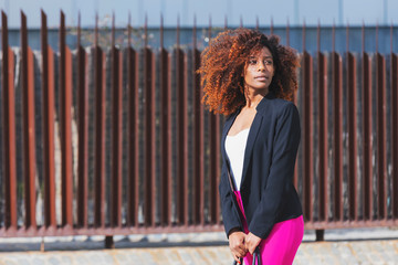 Front view of young beautiful curly woman wearing elegant clothes and handbag while standing in the street in sunny day