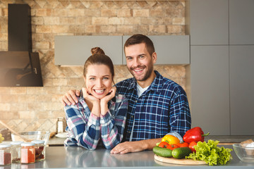 Wall Mural - Young couple at home leaning on table in kitchen together hugging looking camera happy