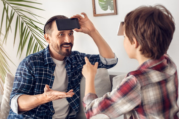 Wall Mural - Father and little son at home sitting on sofa playing heads up with smartphone dad looking confused at boy explaining word