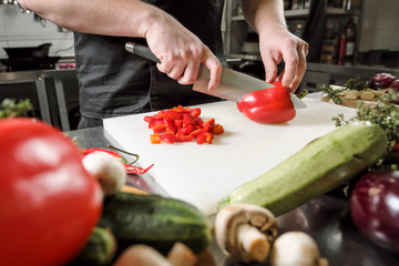 Wall Mural - Male chef cuts vegetables for salad in a restaurant in a black apron. White cutting board, closeup of hands.