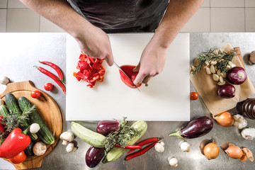 Wall Mural - Male chef cuts vegetables for salad in a restaurant in a black apron. White cutting board, closeup of hands.