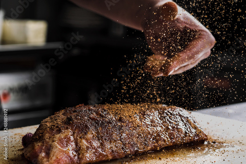 Raw piece of meat, beef ribs. The hand of a male chef puts salt and spices on a dark background.
