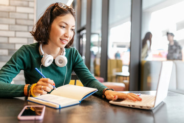 Young happy business asian woman taking notes in notebook and working with laptop pc or computer. Education and working concept. Modern freelance professions