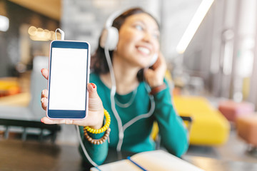 Happy young girl listening to music in headphones and showing blank screen of smartphone with copyspace
