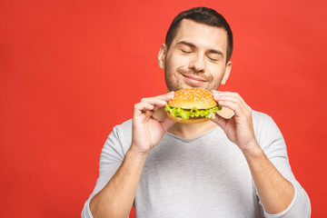 Young man holding a piece of hamburger. Student eats fast food. Burger is not helpful food. Very hungry guy. Diet concept. Isolated over red background.