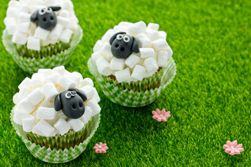 Wall Mural - Easter sheep cupcakes on green grass background, homemade cup cakes shaped funny sheeps with marshmallow and black fondant icing faces, creative treats for kids on Easter holiday