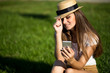 © zadorozhna - Beautiful young woman sitting on grass and using phone in summer park.